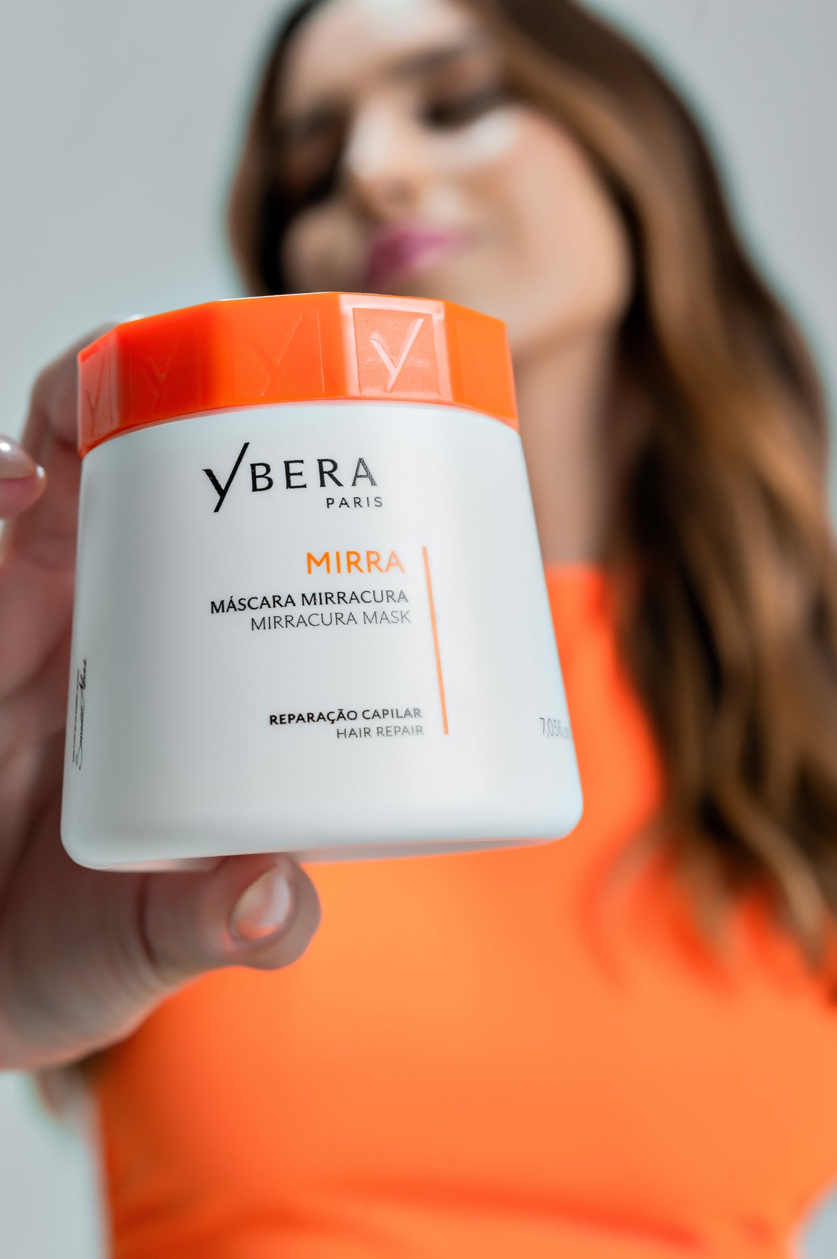 Mujer con camiseta naranja mostrando un tarro blanco de Mirra de Ybera Paris con tapa naranja.-81