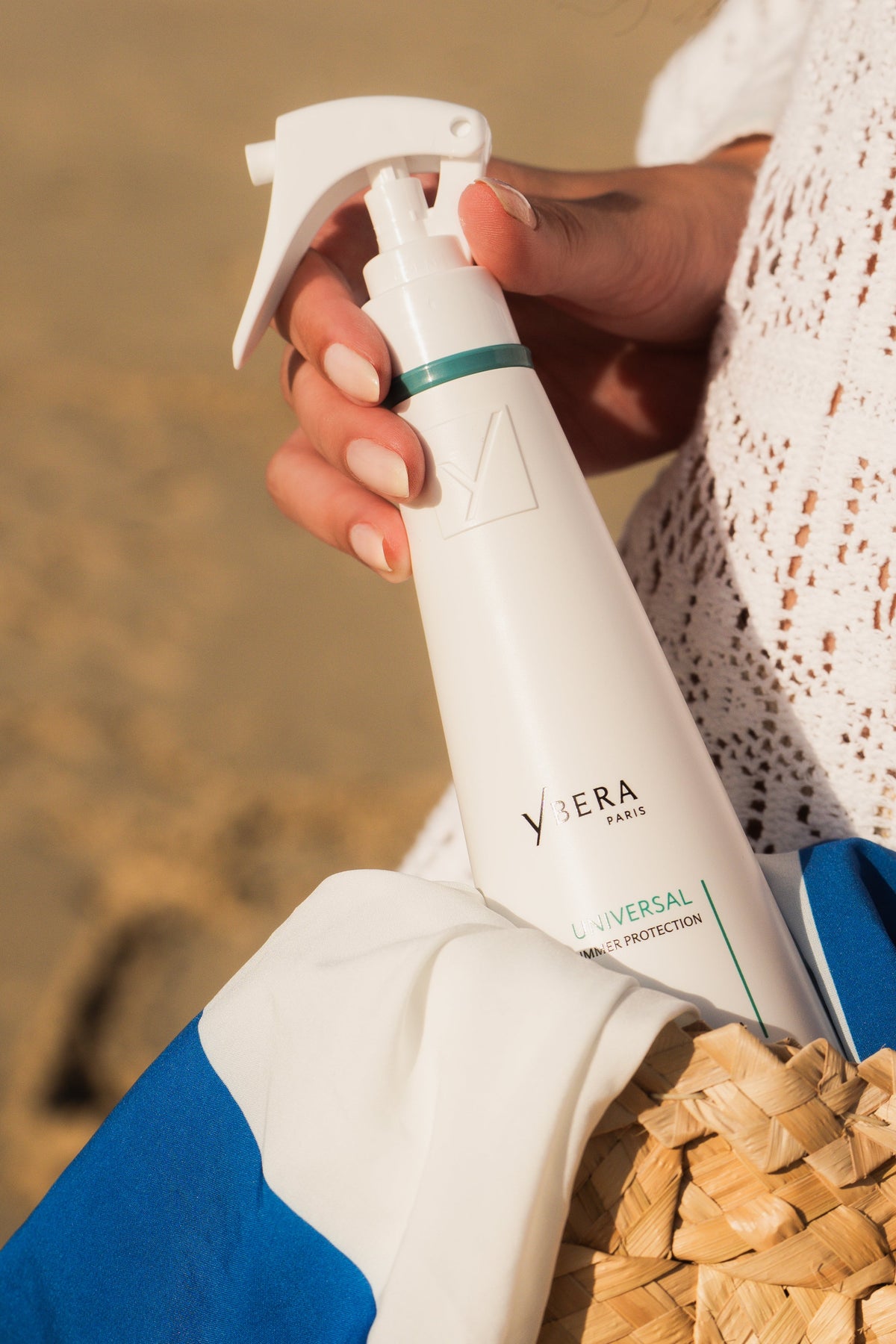 Persona sosteniendo un spray blanco Universal de Ybera Paris con detalles verdes junto a una bolsa de playa.-72