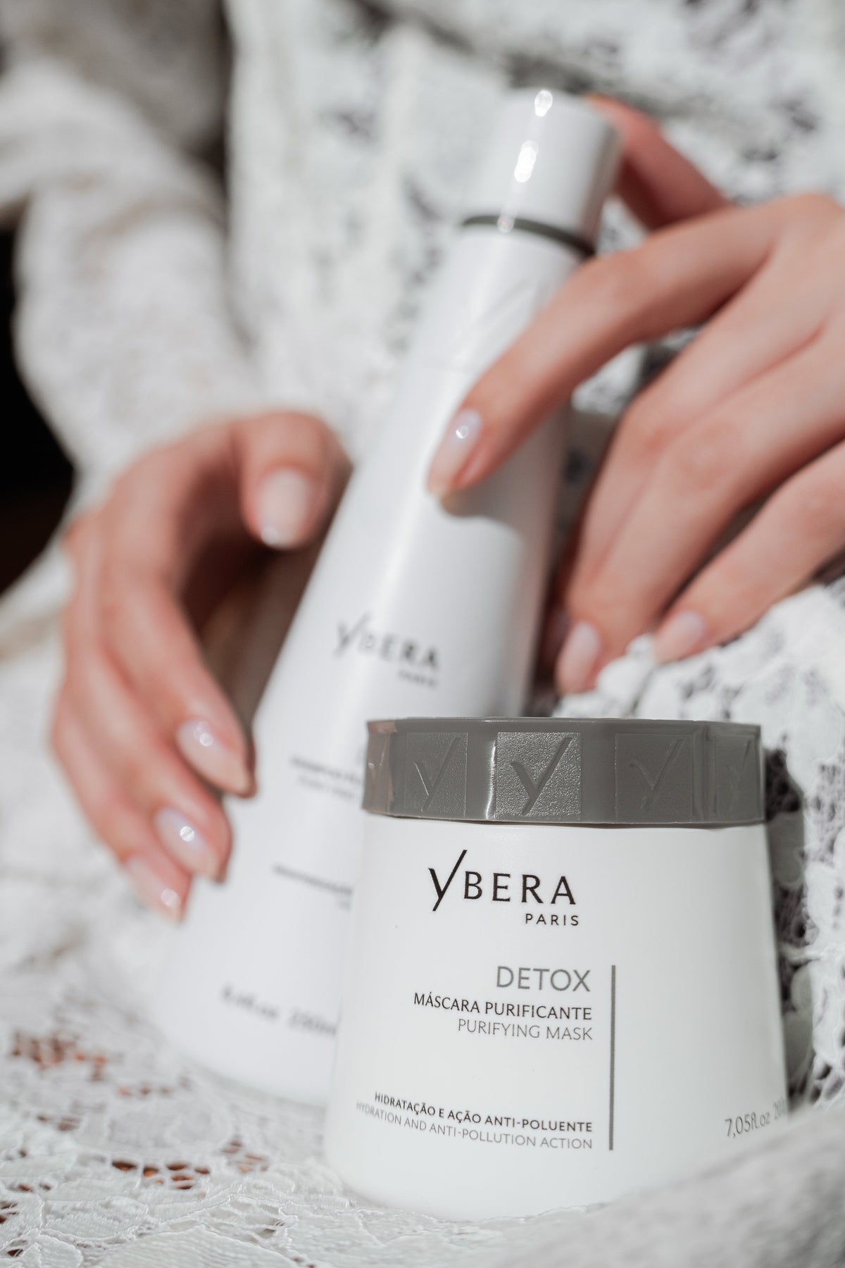 Manos sujetando una botella blanca junto a un tarro de mascarilla purificante Detox de Ybera Paris con tapa gris.-51