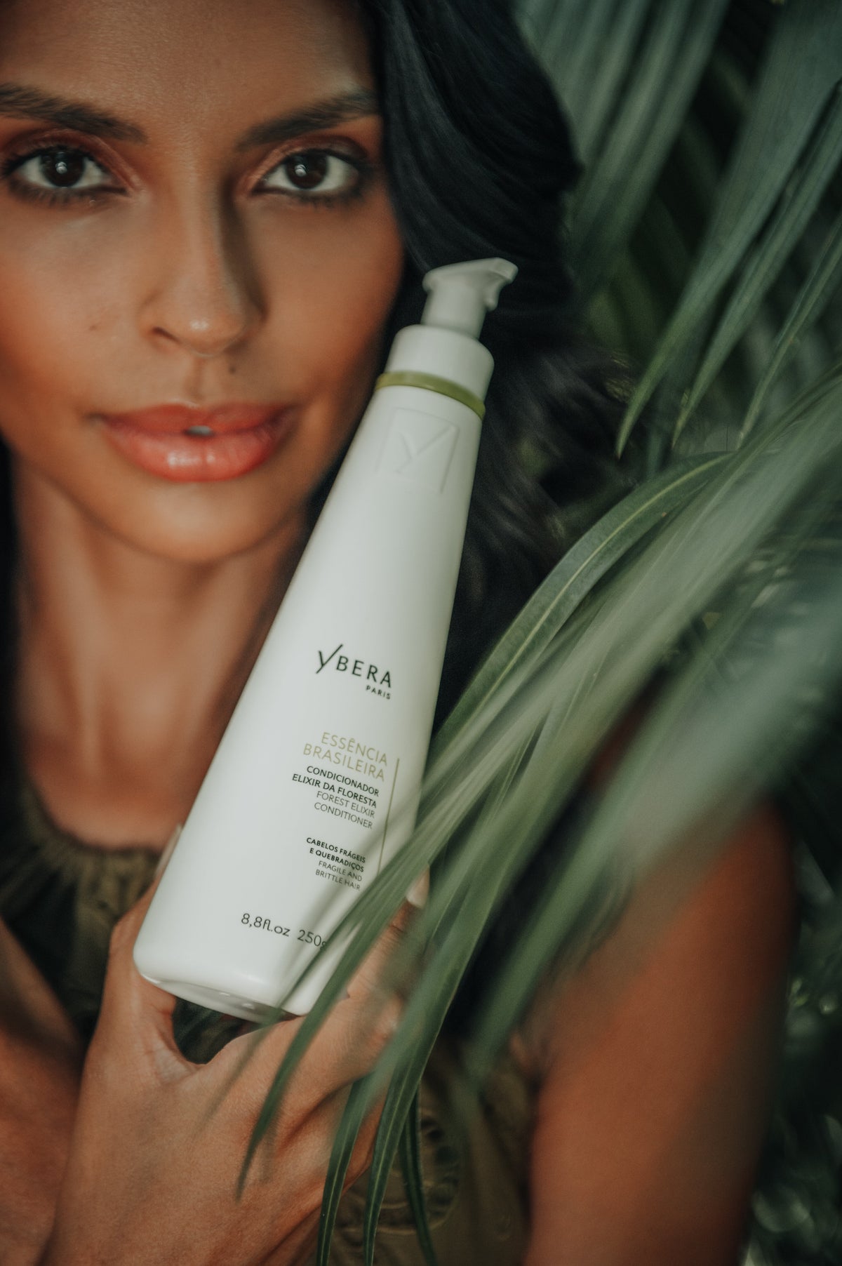 Mujer sosteniendo un envase blanco de acondicionador Essência Brasileira de Ybera Paris junto a hojas verdes.-59